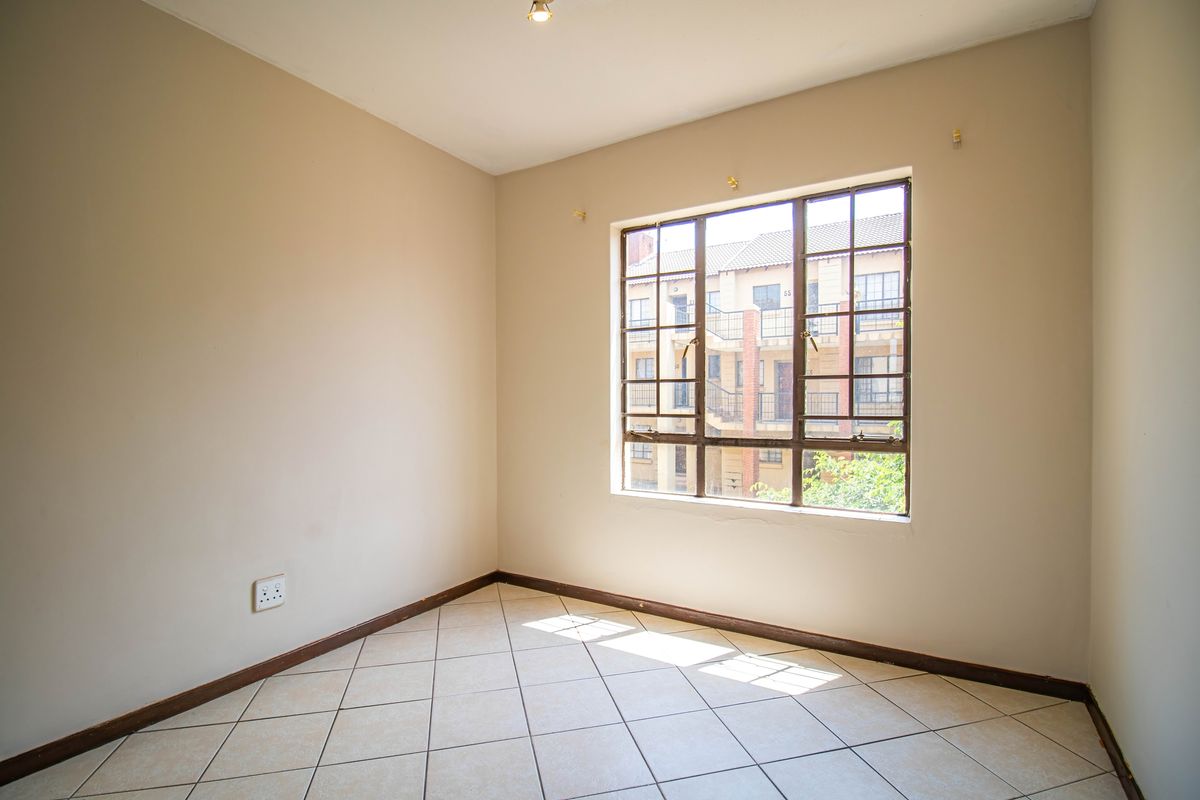 A bright, empty bedroom featuring neutral walls, tiled flooring, and a large steel-framed window overlooking the apartment complex.