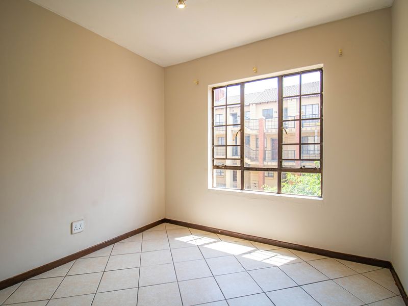 A bright, empty bedroom featuring neutral walls, tiled flooring, and a large steel-framed window overlooking the apartment complex.