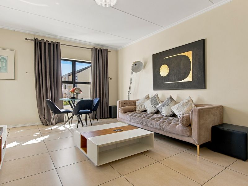 A bright and well-maintained living room featuring neutral tiled flooring, a large window allowing natural light, and a clean, contemporary aesthetic.
