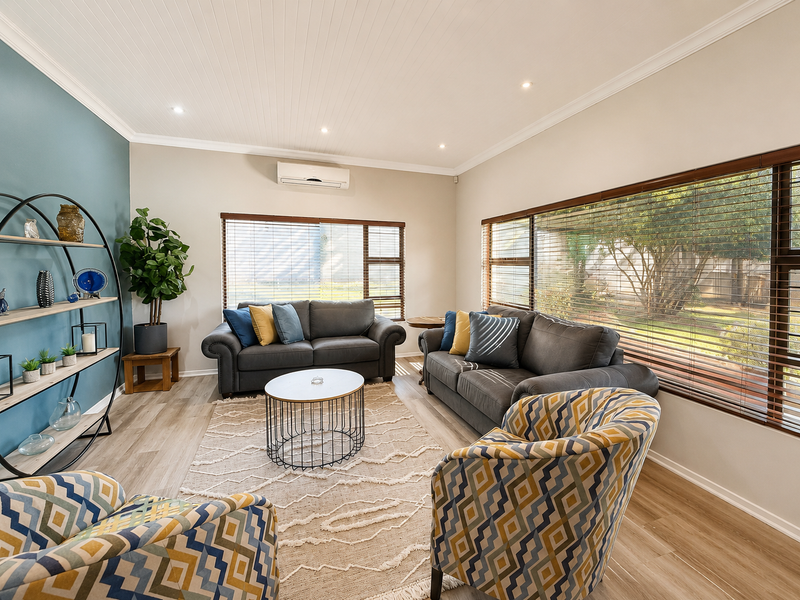A well-maintained living room featuring timber-look flooring, recessed downlighting, split-system air conditioning, and large windows with venetian blinds overlooking the garden.