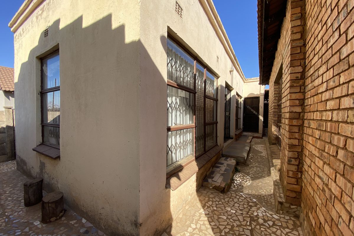 A side exterior view of the property featuring plastered walls, exposed brickwork, metal security gates, and a paved walkway.