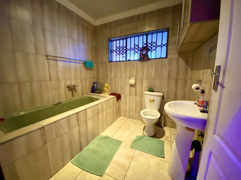 A functional bathroom featuring a built-in bathtub, pedestal sink, toilet, and neutral wall and floor tiling with security bars on the window.