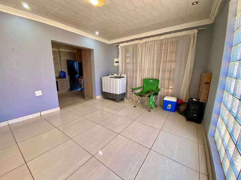 A spacious living area featuring large floor tiles, decorative ceiling cornices, and a unique glass block feature wall, leading into an adjacent kitchen space.
