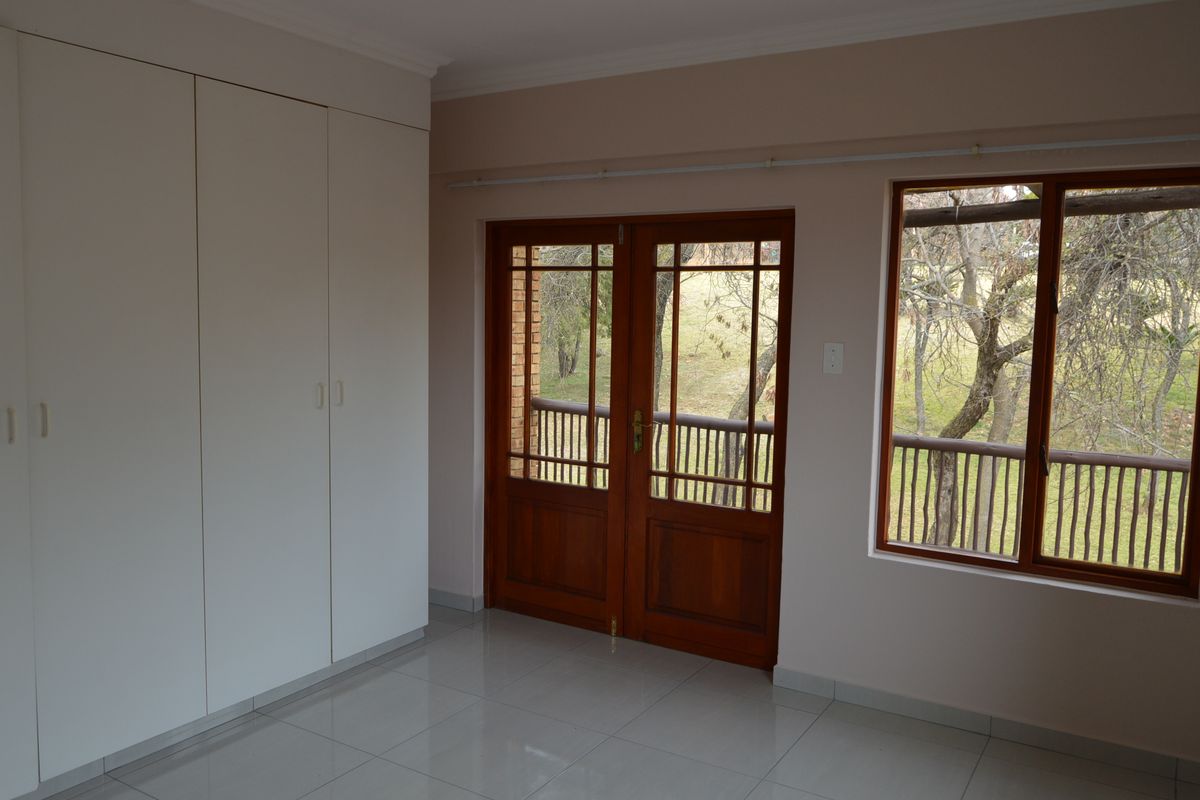 A bright, empty bedroom featuring tiled flooring, built-in cupboards, and wooden framed doors leading to an outdoor balcony area.