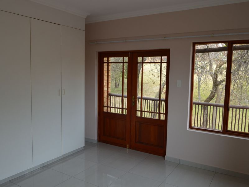 A bright, empty bedroom featuring tiled flooring, built-in cupboards, and wooden framed doors leading to an outdoor balcony area.