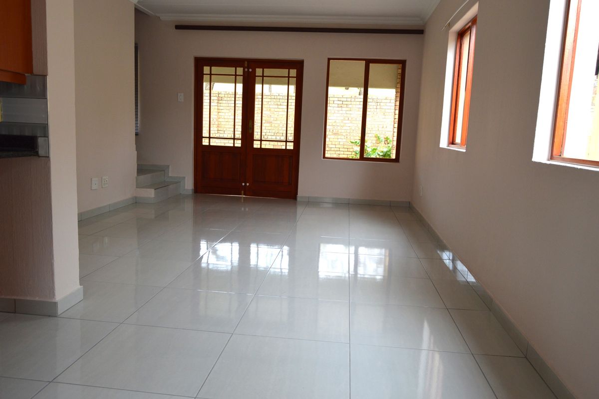 A bright, empty living area featuring polished tiled flooring, wooden framed windows, and double doors leading to the exterior.