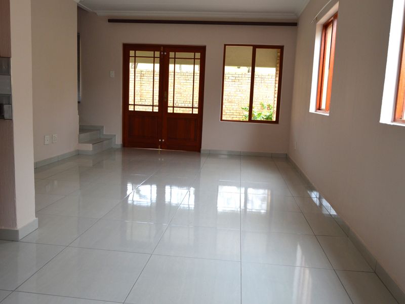 A bright, empty living area featuring polished tiled flooring, wooden framed windows, and double doors leading to the exterior.