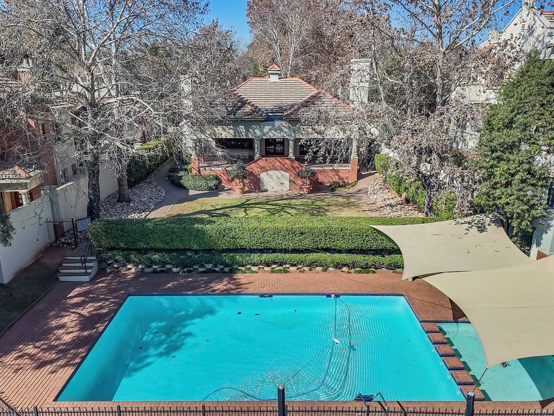 An elevated view of a well-maintained swimming pool area featuring shade sails, brick paving, and a landscaped garden leading to the main residence.