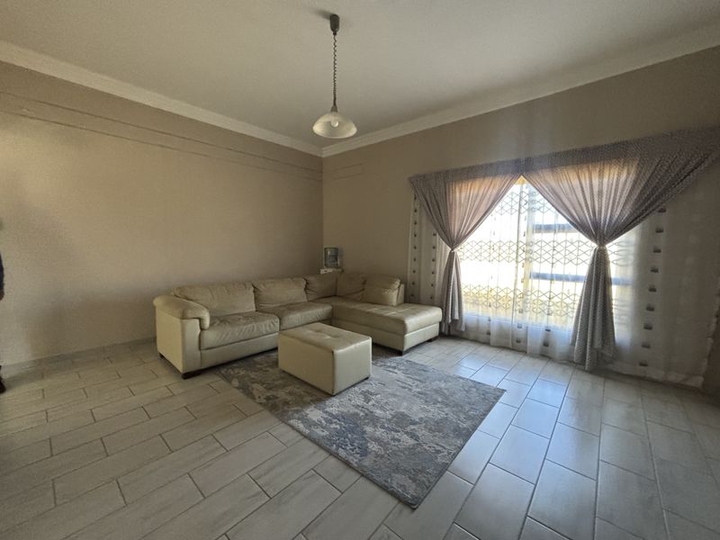 A spacious living room featuring neutral wall tones, tiled flooring, and a large window with security gate, providing a bright and functional living space.