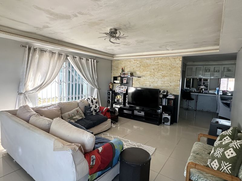 Open plan living area featuring a stone feature wall and tiled flooring, with an adjacent kitchen area. Ceiling shows signs of water damage.