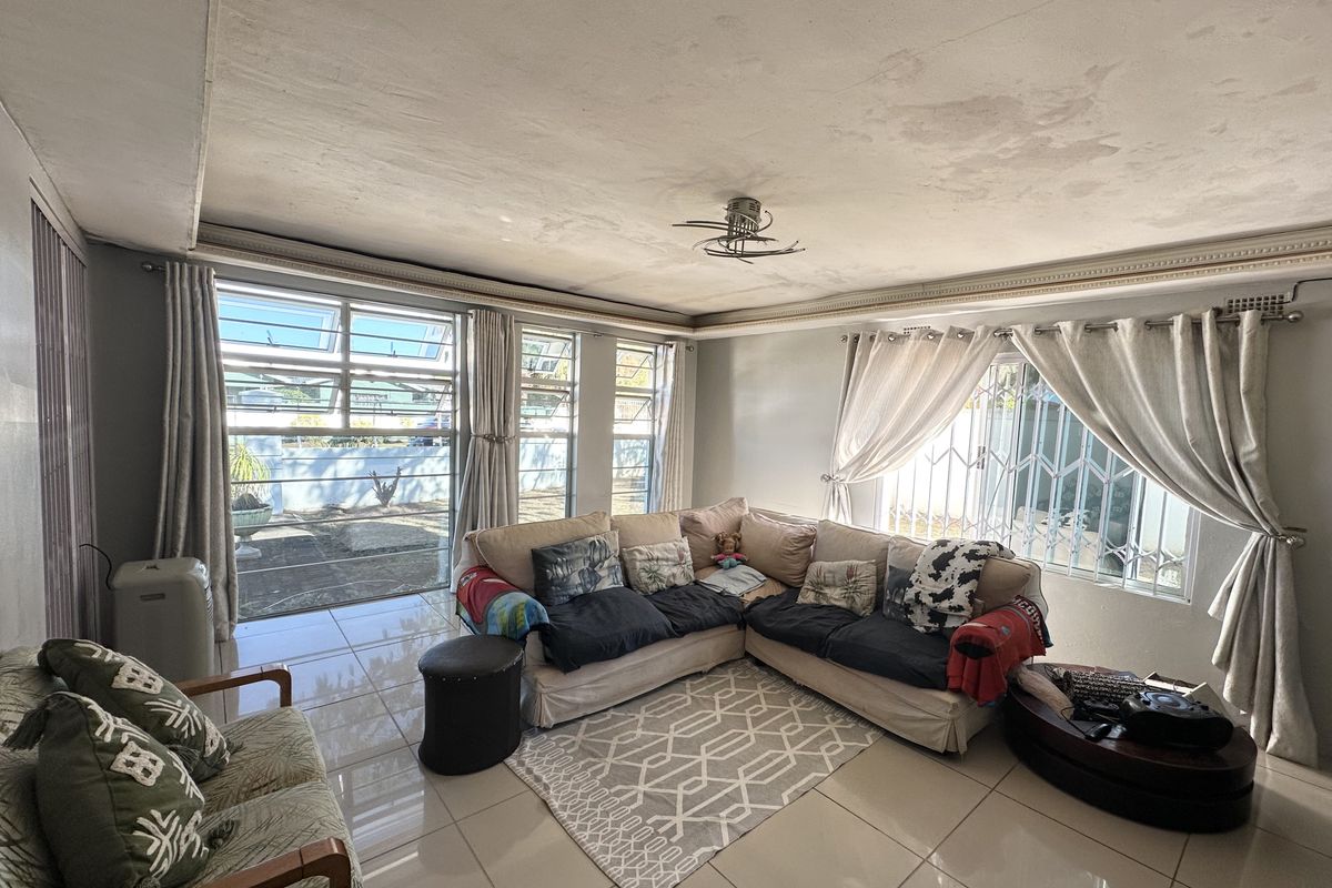 Spacious living room featuring tiled flooring, large windows with security gates, and a ceiling fan. The ceiling shows signs of water damage and wear.