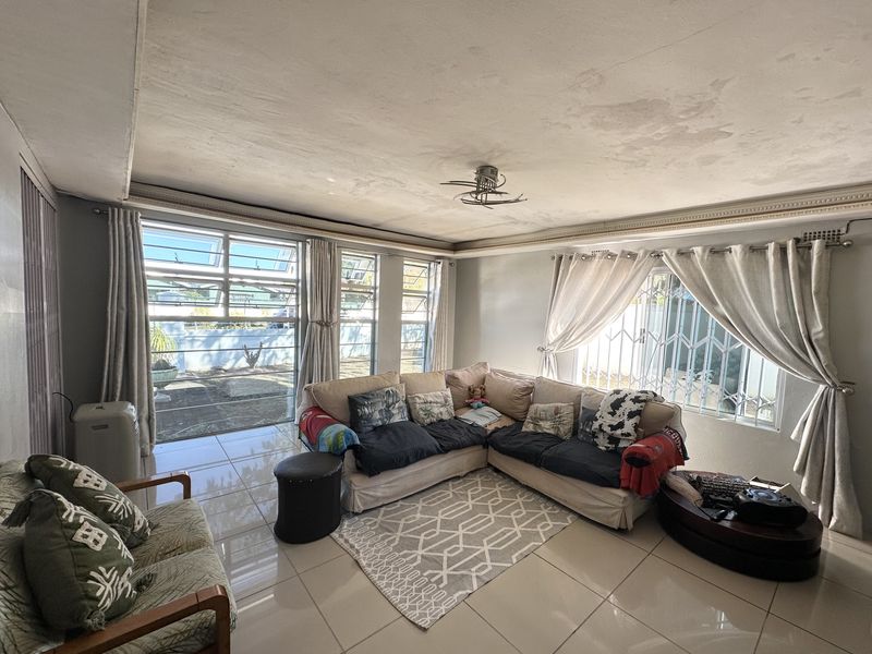 Spacious living room featuring tiled flooring, large windows with security gates, and a ceiling fan. The ceiling shows signs of water damage and wear.