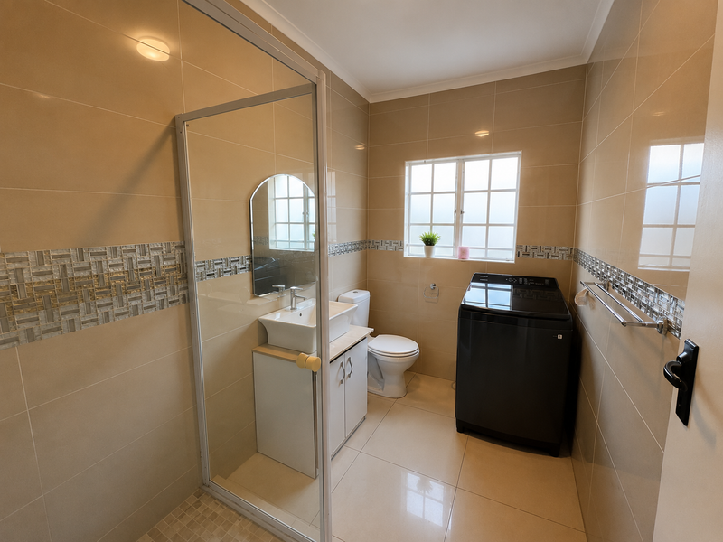 A functional bathroom featuring a glass-enclosed shower, vanity with basin, toilet, and neutral tiled walls with a decorative mosaic border.