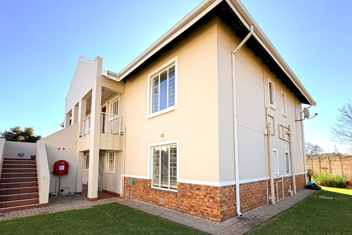 Rear view of a two-story townhouse featuring a brick-base facade, external staircase, balcony, and well-maintained surrounding lawn and paved pathways.
