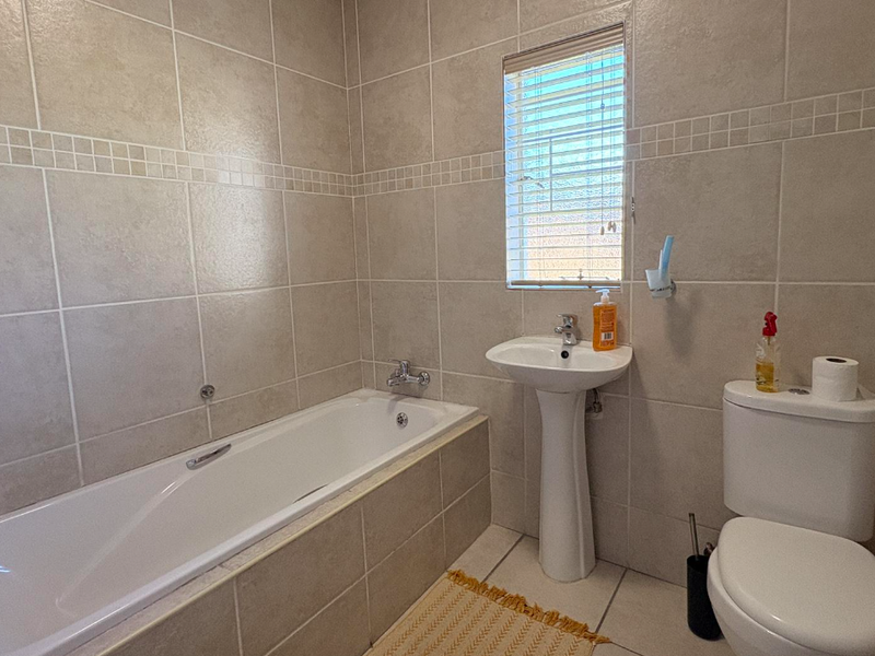 A functional bathroom featuring a built-in bathtub, pedestal basin, and toilet, finished with neutral wall and floor tiling.