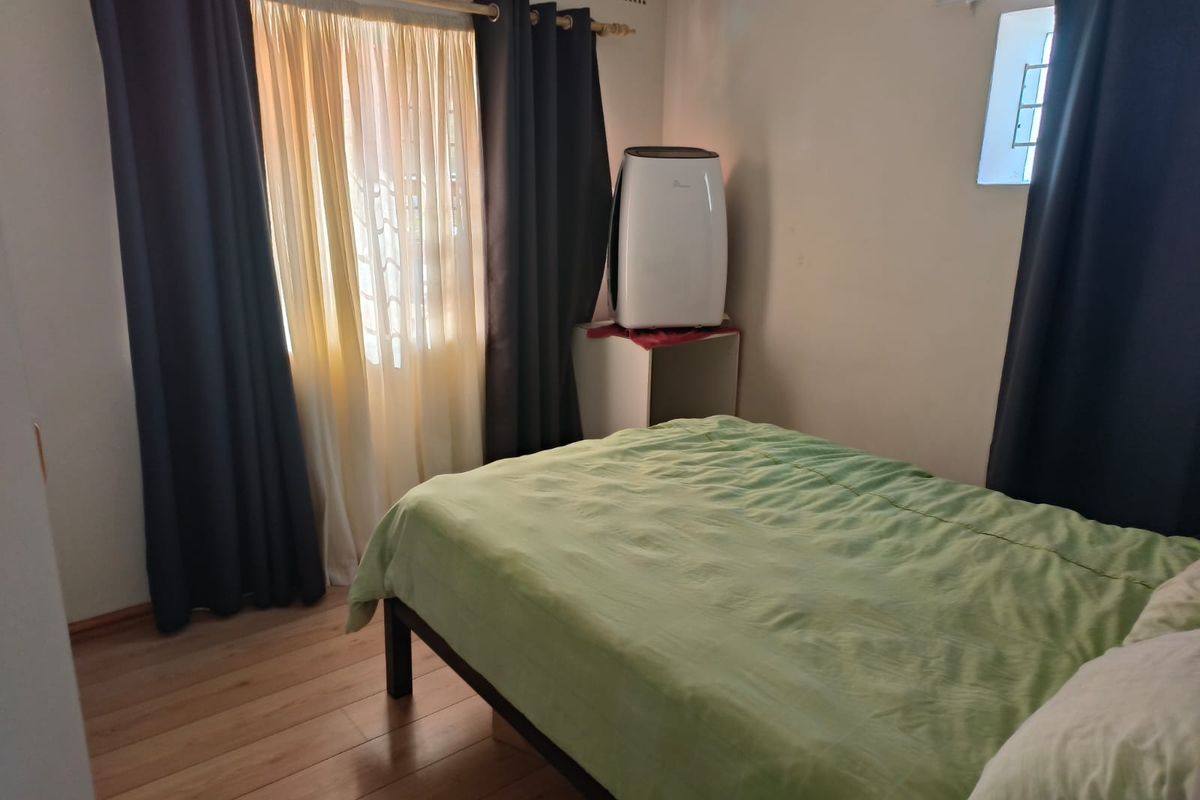 A functional bedroom featuring laminate flooring, natural light through a window with security bars, and a built-in ventilation grille.