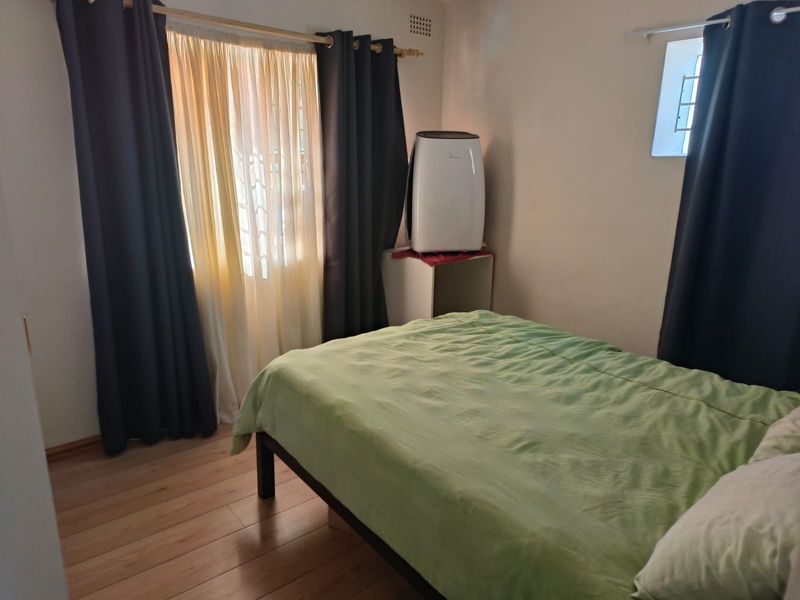A functional bedroom featuring laminate flooring, natural light through a window with security bars, and a built-in ventilation grille.