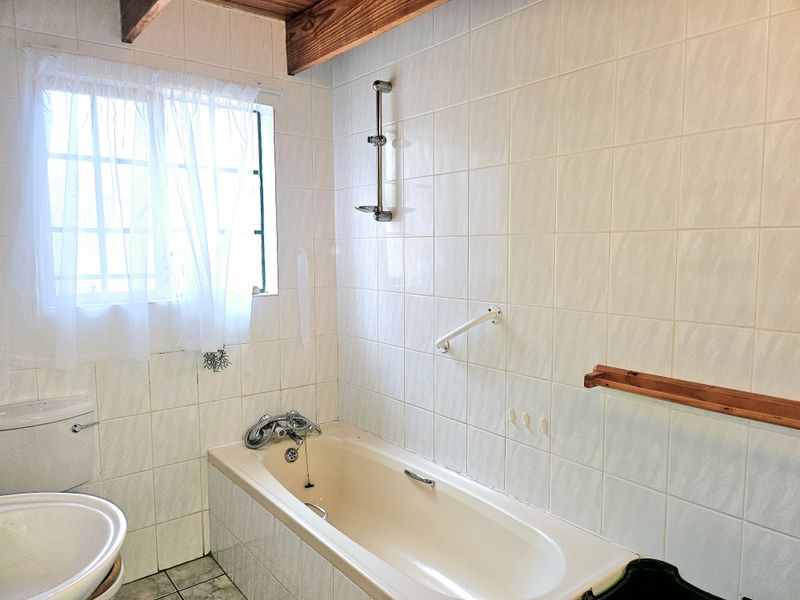 A dated bathroom featuring tiled walls, a bathtub with a shower riser rail, a toilet, and exposed timber ceiling beams.