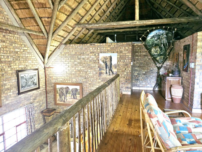 A rustic mezzanine hallway featuring exposed timber roof trusses, a thatched ceiling, facebrick walls, and a timber balustrade overlooking the lower level.