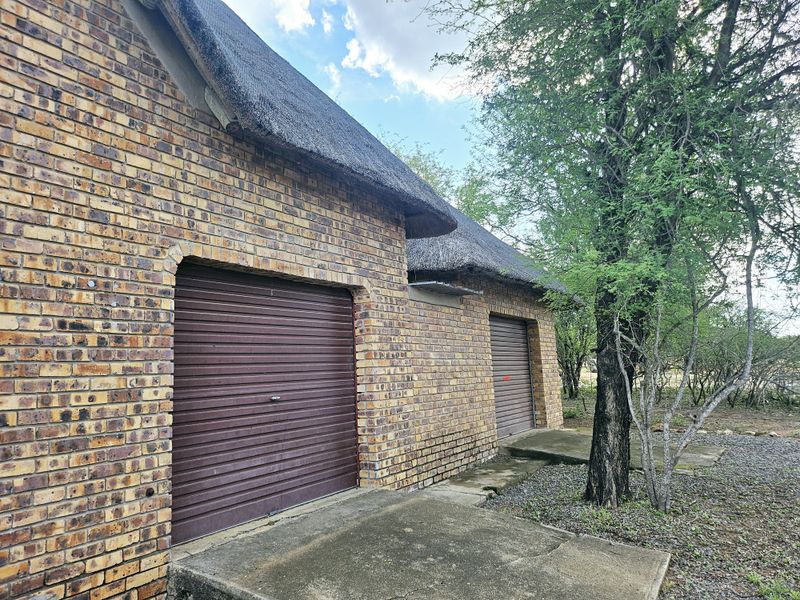 A detached face brick garage building featuring a traditional thatch roof and two roller shutter doors, set within a natural garden environment.