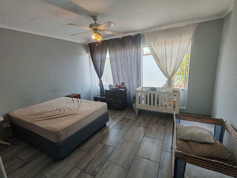 A spacious bedroom featuring modern wood-look floor tiles, a ceiling fan, and large windows that provide ample natural light.