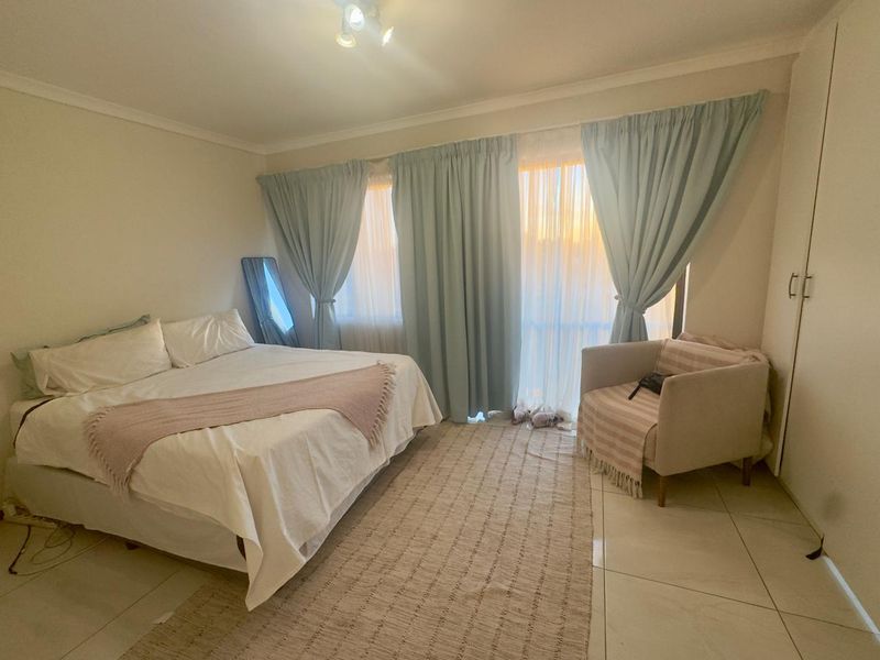 A well-lit bedroom featuring neutral tiled flooring, a built-in wardrobe, and large windows with curtains, providing a comfortable and functional living space.