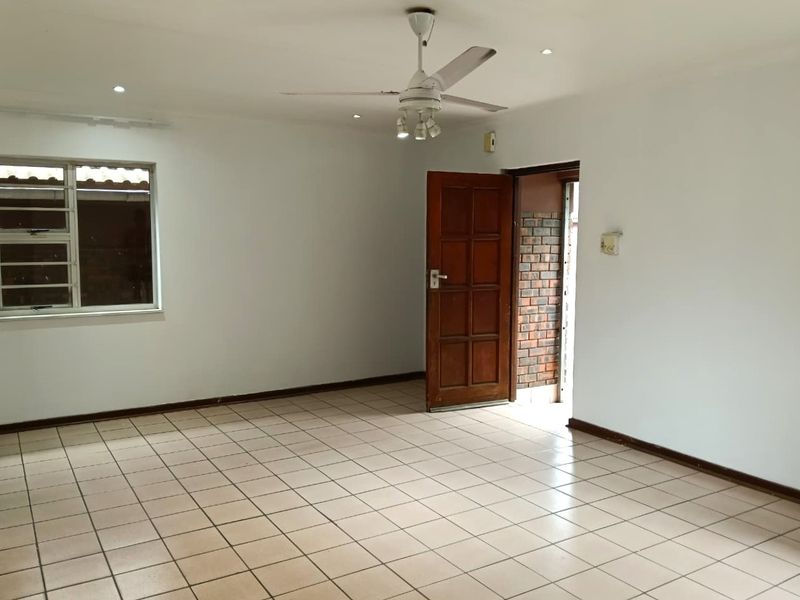An empty living room featuring neutral tiled flooring, a ceiling fan, recessed lighting, and a timber entrance door leading to an exterior brick walkway.