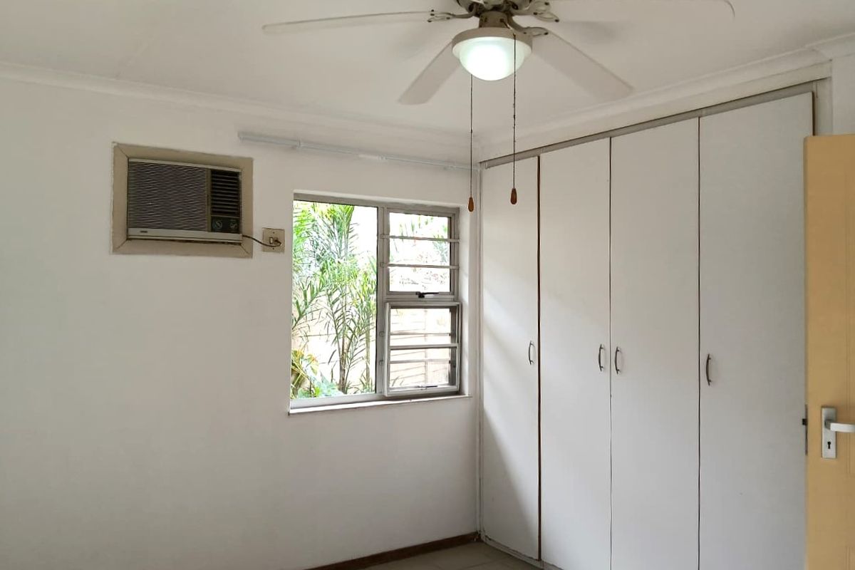 A functional bedroom featuring built-in cupboards, a ceiling fan, and a window-mounted air conditioning unit, offering a blank canvas for personalization.