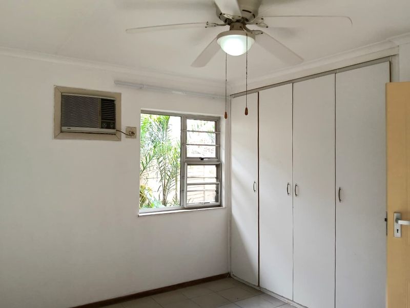 A functional bedroom featuring built-in cupboards, a ceiling fan, and a window-mounted air conditioning unit, offering a blank canvas for personalization.