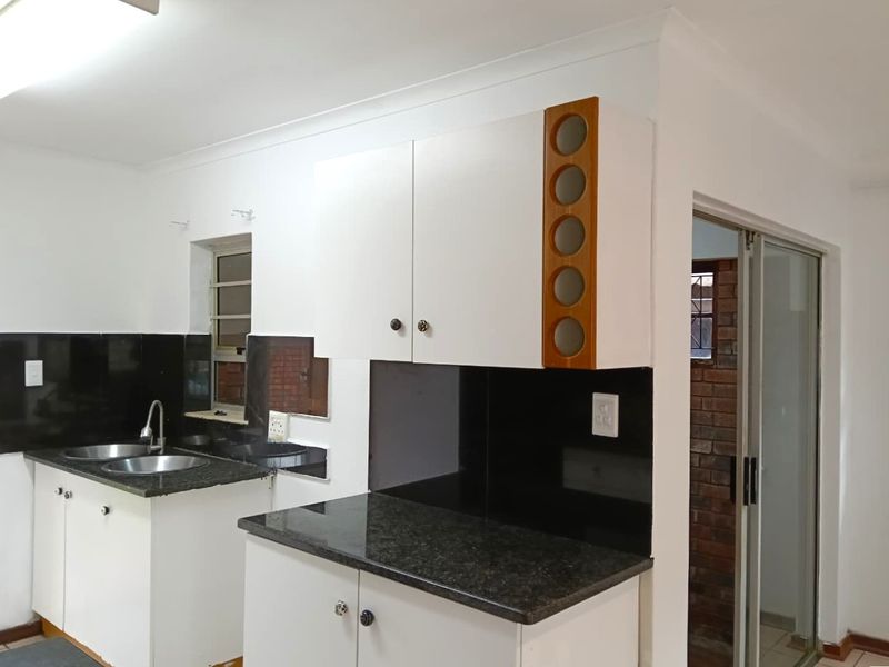A functional kitchen featuring white cabinetry, dark granite countertops, a double bowl sink, and a black tiled backsplash, leading to an adjacent area.