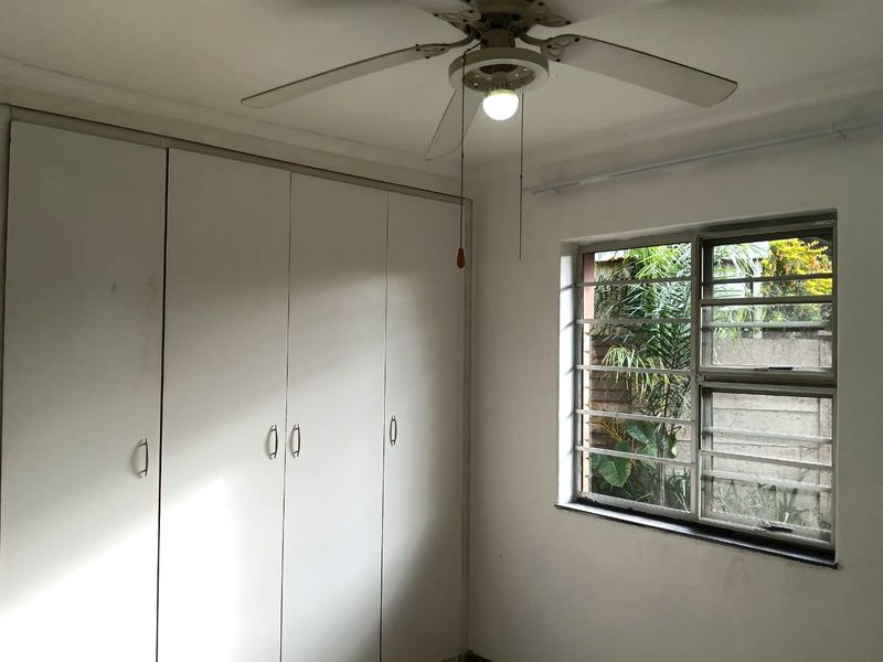 A simple bedroom featuring built-in cupboards, a ceiling fan, and security bars on the window, offering a functional space ready for personalization.
