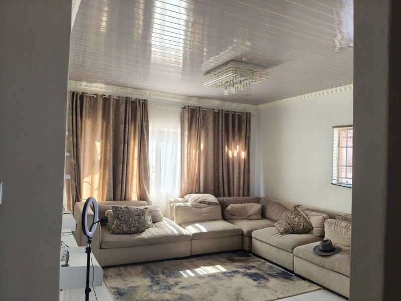 A comfortable living room featuring a glossy panelled ceiling, decorative cornice, and a central crystal light fixture, providing a bright and inviting space.