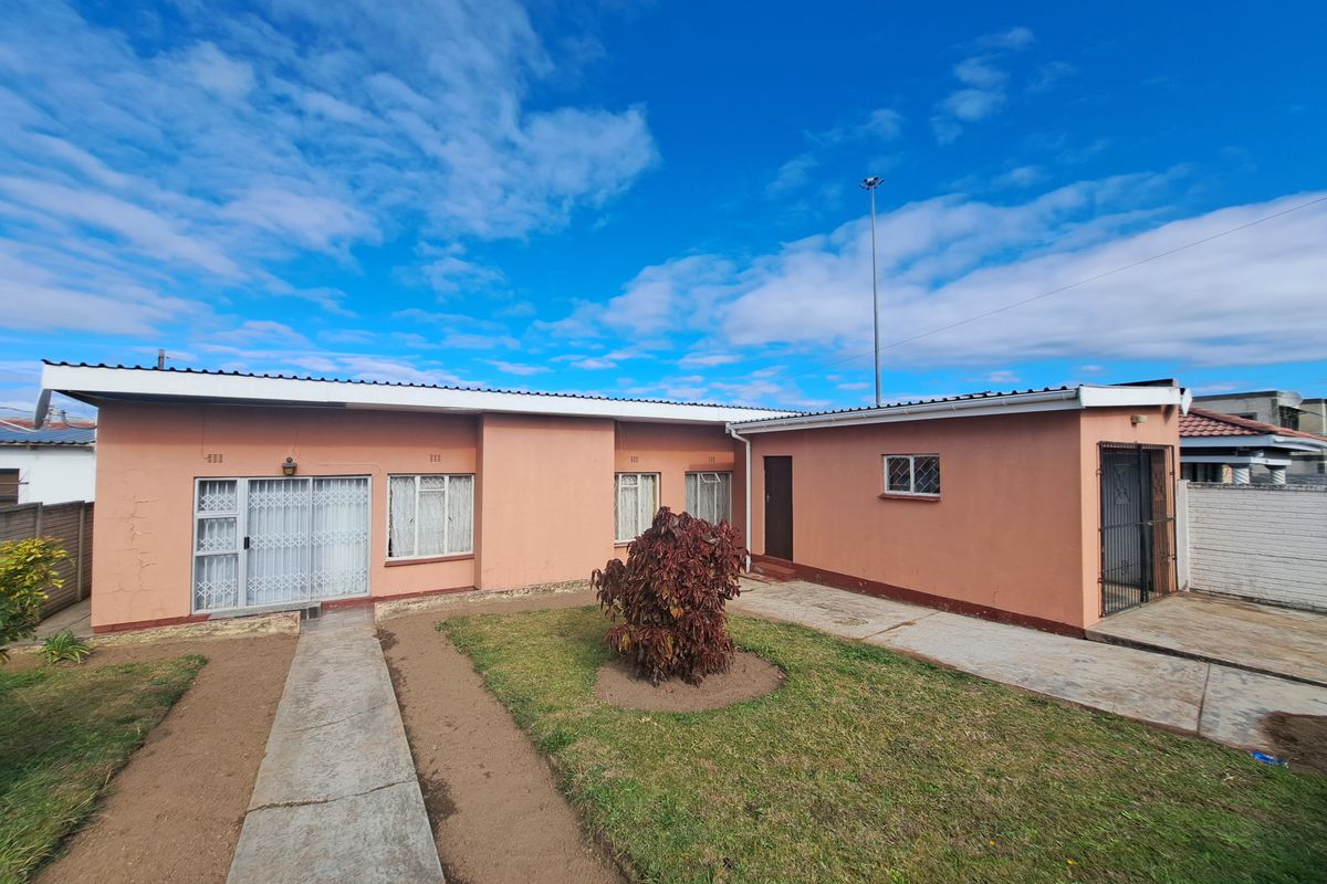 Rear view of a single-story house featuring a concrete walkway, security gates, and a small garden area with exterior wall plaster showing signs of wear.