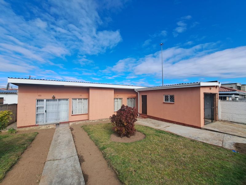 Rear view of a single-story house featuring a concrete walkway, security gates, and a small garden area with exterior wall plaster showing signs of wear.