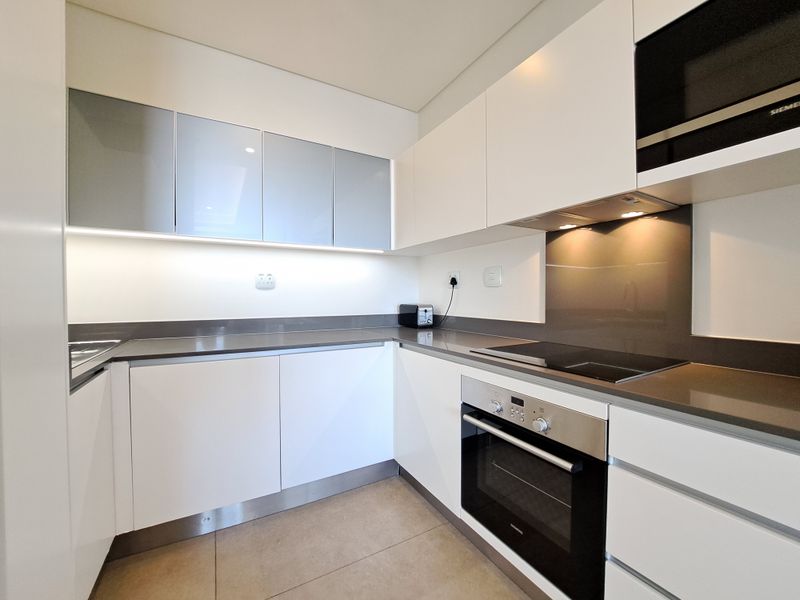 Modern kitchen featuring sleek white cabinetry, stone countertops, integrated appliances including an oven, hob, and microwave, complemented by under-cabinet lighting and tiled flooring.