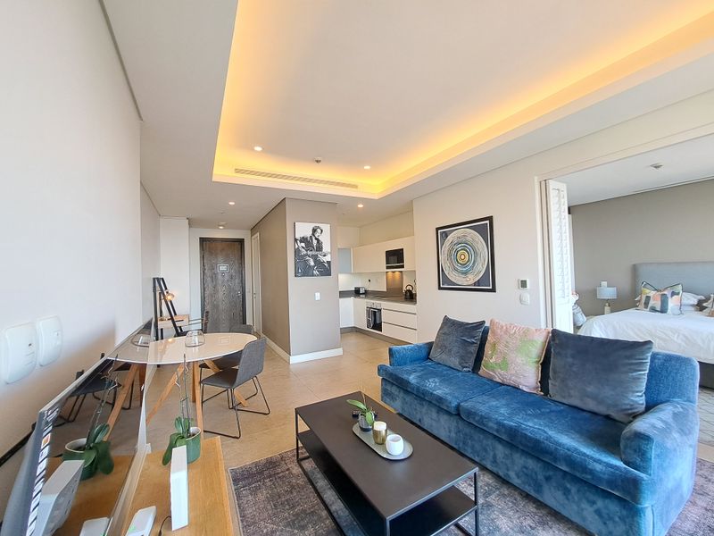 Modern open-plan living area featuring recessed cove lighting, sleek kitchen cabinetry with integrated appliances, and tiled flooring, leading into a separate bedroom space.