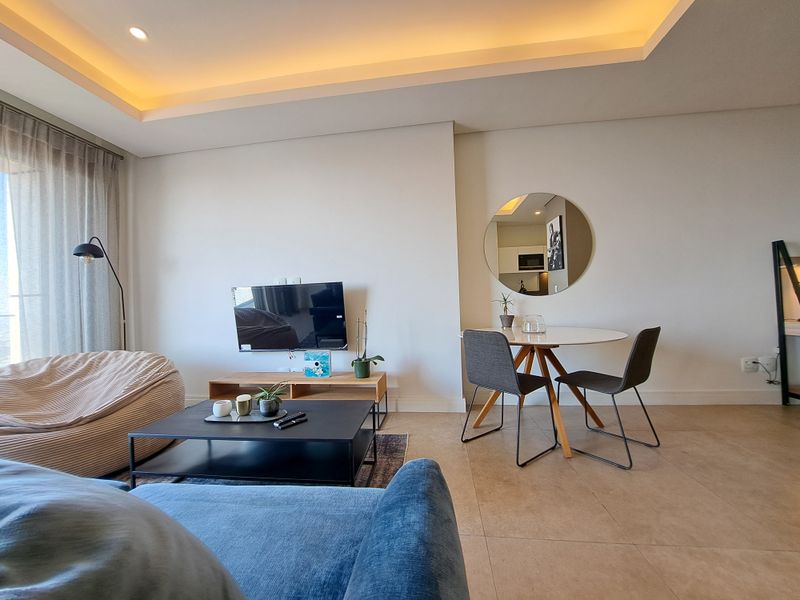 Modern open-plan living area featuring tiled floors, recessed ceiling lighting with cove accents, and a bright, neutral aesthetic with access to a balcony.