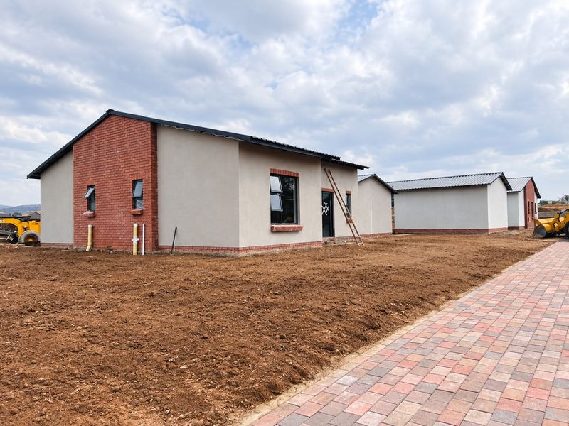 A newly constructed residential property featuring a combination of brick and plastered exterior walls, metal roofing, and a paved walkway, currently undergoing final site development.