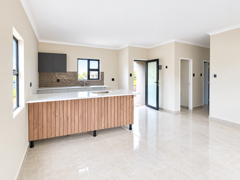 A bright, modern open-plan kitchen featuring a wood-paneled island, stone countertops, tiled backsplash, and neutral ceramic flooring in a clean, vacant space.