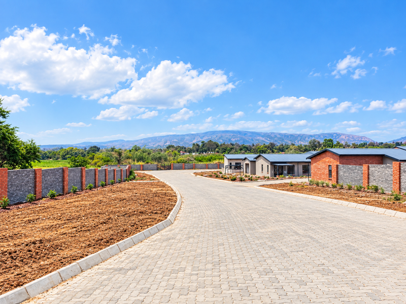 A view of a paved residential driveway leading into a modern estate, featuring brick boundary walls and scenic mountain views in the background.