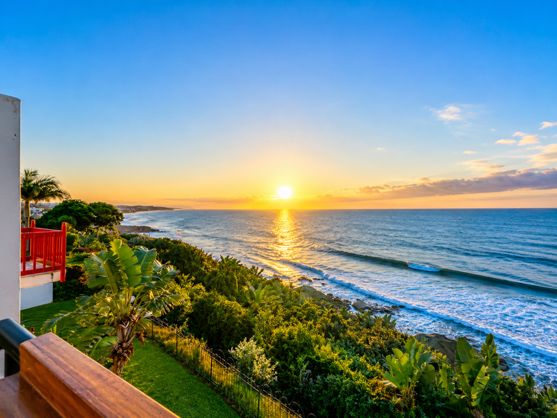 A stunning oceanfront view at sunset, captured from a balcony overlooking the coastline, lush greenery, and the sea.