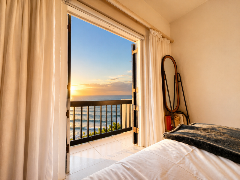 A bright bedroom featuring a direct walk-out to a private balcony with stunning ocean views and sunset exposure.