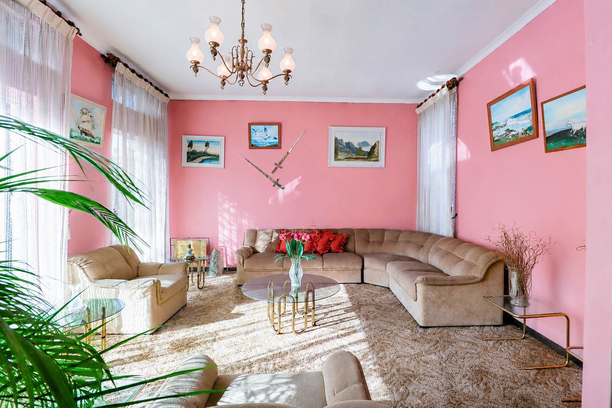 A spacious living room featuring a coved ceiling, chandelier lighting, and large windows with curtain rails, currently finished with dated wall colors and carpeting.
