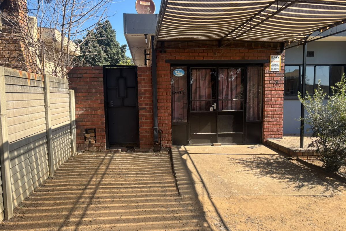 Rear exterior area featuring a brick facade, metal security gate, and a covered patio space with concrete paving.
