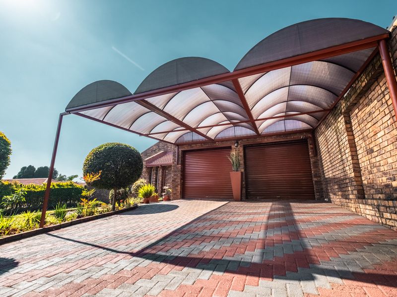 A well-maintained double garage featuring roll-up doors, a paved driveway, and a modern curved polycarbonate carport structure.