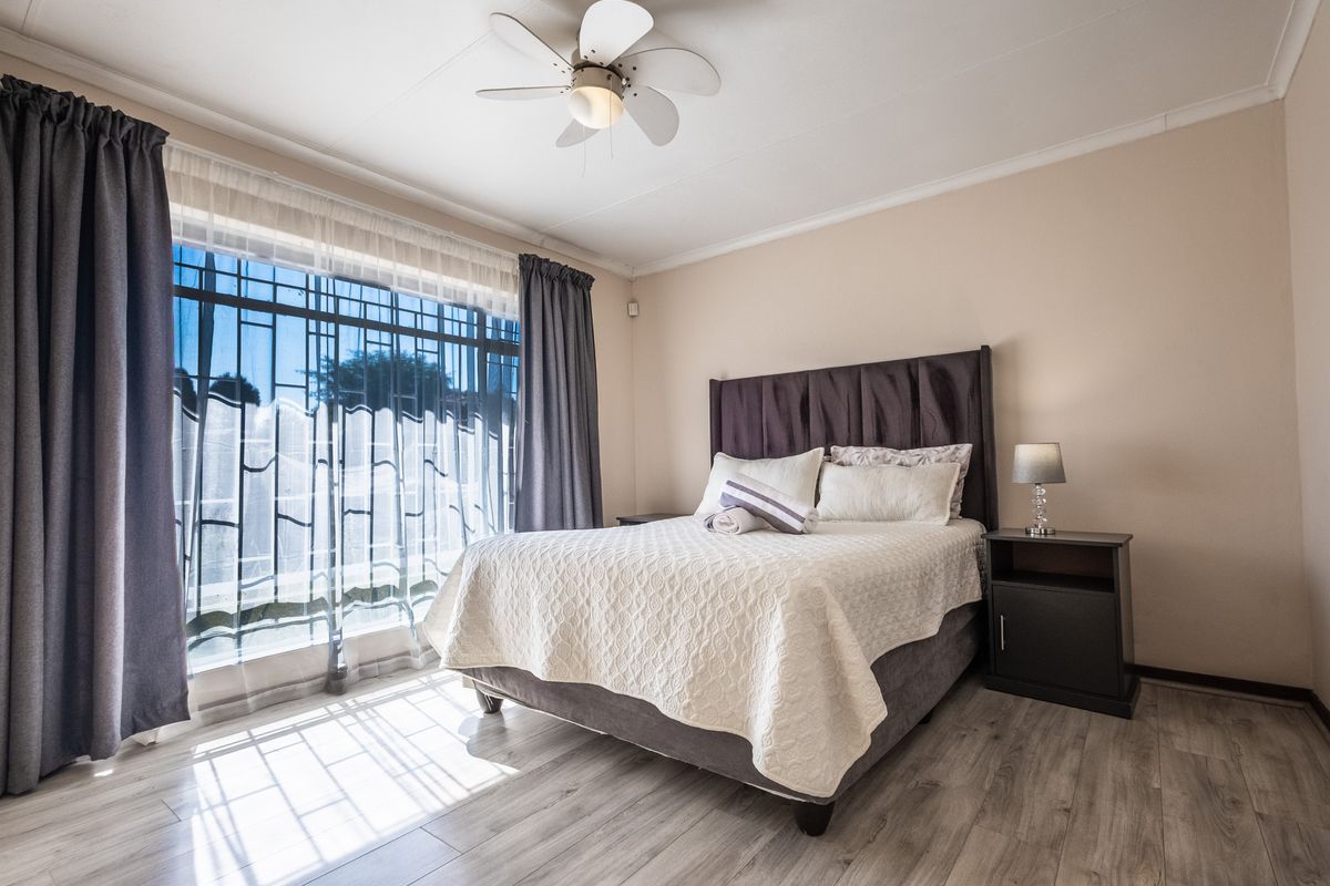 A well-maintained bedroom featuring light-toned laminate flooring, a ceiling fan, and large windows fitted with security bars.