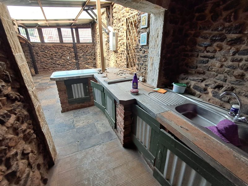Covered outdoor patio area featuring rustic stone walls, built-in brick cabinetry with a stainless steel sink, and durable tiled flooring.