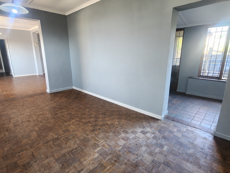 An empty living area featuring classic parquet flooring, neutral grey walls, and direct access to adjacent rooms and a tiled entryway.