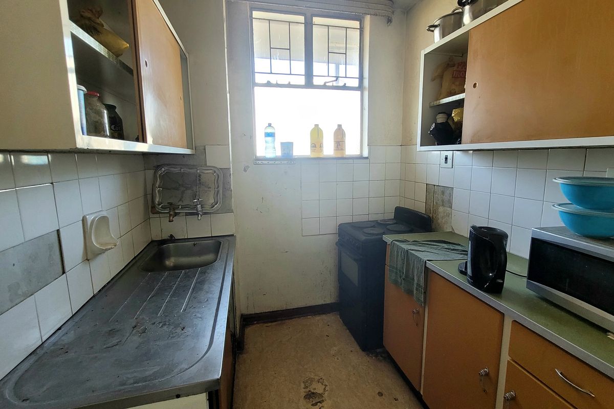 A compact, dated kitchen featuring original cabinetry, a stainless steel sink, and a freestanding stove, requiring modernization and cosmetic updates.