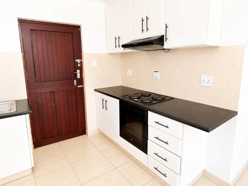 A clean, functional kitchen featuring white cabinetry, dark countertops, a built-in oven with hob, extractor fan, and neutral tiled finishes.